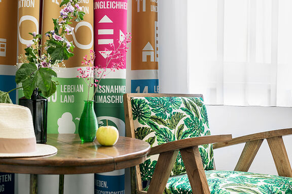 Ein Holzsessel mit einer Polsterung im Pflanzenmuster steht neben einem Holztisch auf dem zwei Vasen mit Blumen stehen, im Hintergrund ist ein Vorhand mit den Sustainable Development Goals als Muster