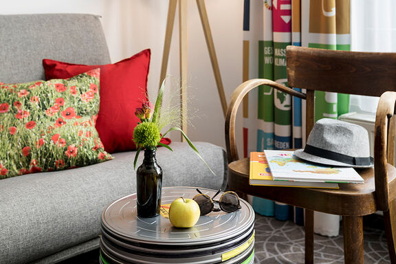 A gray sofa stands next to a coffee table with a vase of flowers, sunglasses and an apple on it
