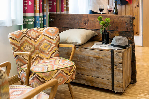 Ein bunter Sessel steht neben einer Holzkommode auf dem ein grauer Hut liegt und auf dem zwei Weingläser mit Rotwein stehen