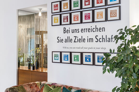 Mehrere Bilder mit den verschiedenen Sustainable Development Goals hängen auf einer weißen Wand, neben der Aufschrift 'Bei uns erreichen Sie alle Ziele im Schlaf'